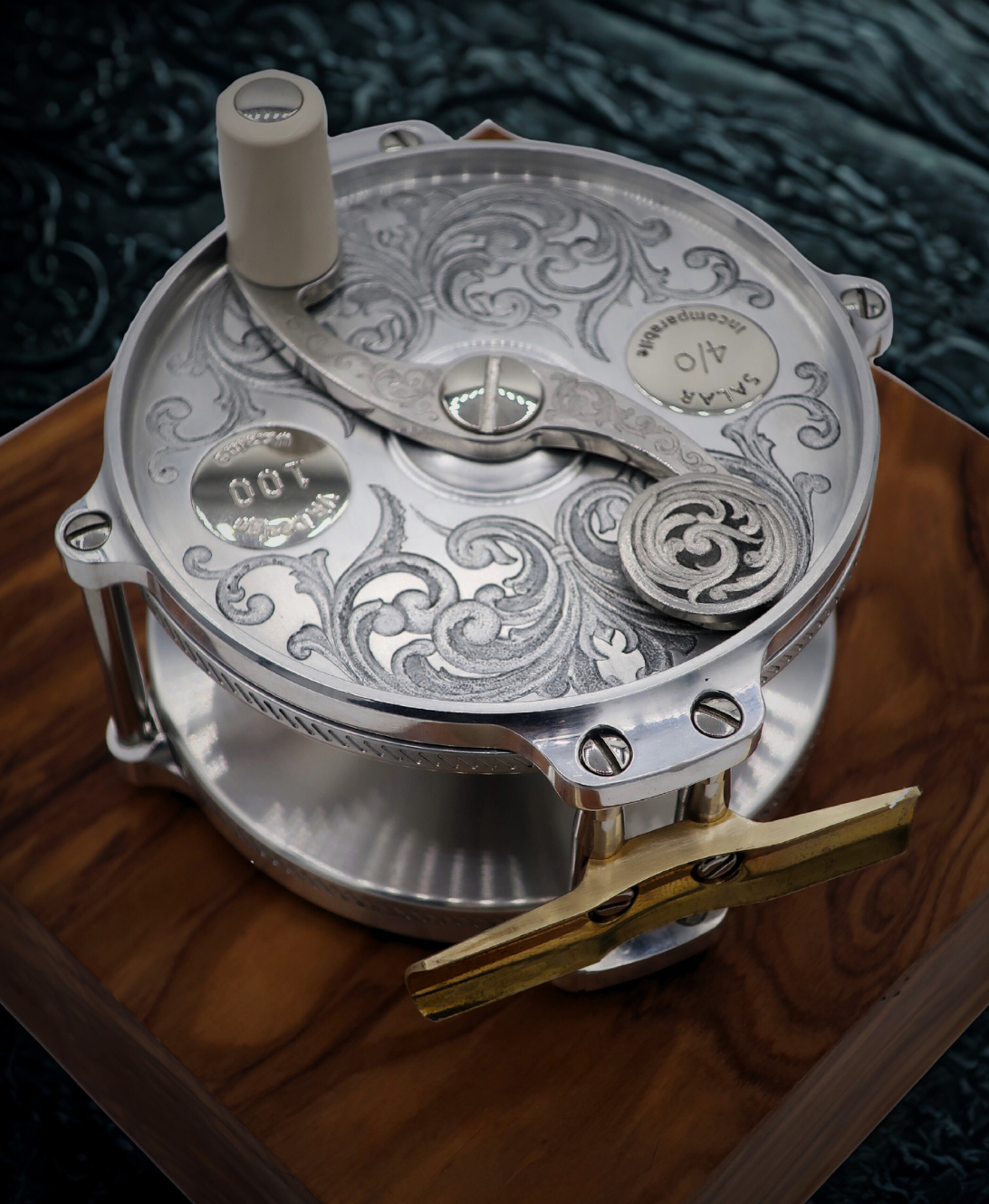 Hand engraved salar fly fishing reel by VRDesigns. The engraving features scrollwork. The reel is sitting on a piece of wood.