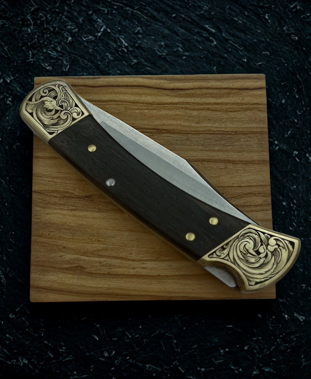 Hand engraved buck 110 folding knife. The engraving features scrollwork. The knife is resting on a square piece of wood.