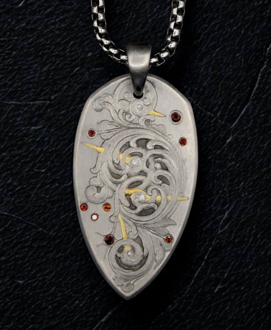 Hand engraved white metal pendant in the shape of a spade. The pendant features scrollwork in the middle, with gold inlay lines. The piece features garnets and diamonds scattered. The piece is on a black background