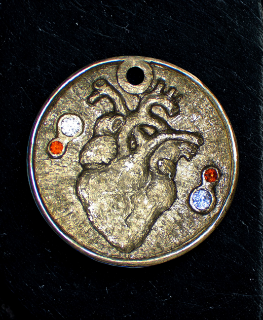 Gold disc pendant featuring a hand engraved life-like heart in the center. The heart has a white sapphire and red garnet on the left and right side.